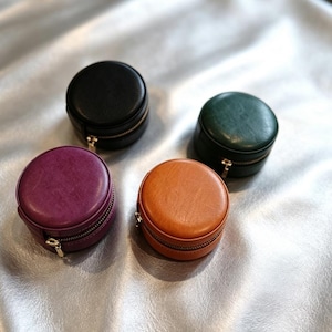 May include: Four small, round, zippered cases in black, dark green, purple, and orange. Each case features a smooth, solid-colored exterior with a matching zipper closure and a small metal pull tab, designed for compact storage of small items.