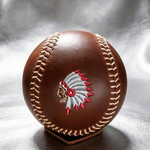 Op de afbeelding: Een bruine leren honkbal met witte stiksels en een centrale geborduurde afbeelding van een persoon met een traditionele hoofdtooi van witte en rode veren. De bal is glad, rond en heeft een gepolijste textuur op het oppervlak.