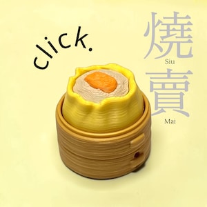 Könnte beinhalten: Eine gelbe und hellbraune Knödel-Figur in einem Bambusdämpfer. Der Text "click." steht links und die chinesischen Schriftzeichen für "Siu Mai" stehen rechts. Der Knödel hat eine texturierte Oberfläche mit einem orangefarbenen Mittelstück.