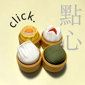 Könnte beinhalten: Vier Miniatur-Dim-Sum-Artikel in kleinen Bambusdampfgarern. Ein gelbes Shumai mit orangefarbener Mitte, ein weißes Brötchen mit roter Oberseite, ein weißer gefalteter Knödel und ein grüner eingewickelter Knödel. Der Text "click." und chinesische Schriftzeichen sind zu sehen.