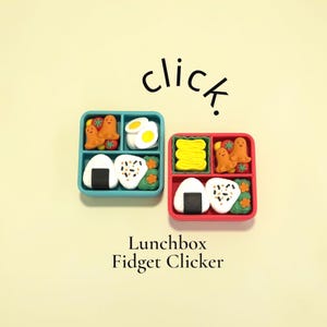 May include: Two square lunchbox fidget clickers in teal and red, each containing miniature food items like onigiri, octopus-shaped sausages, and egg slices. The text 'click.' appears above, and 'Lunchbox Fidget Clicker' is printed below the items.