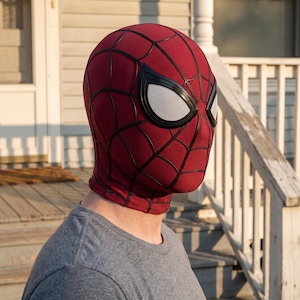 May include: A red fabric mask with a black web pattern and large white mesh eye lenses. The mask covers the entire head and neck, featuring a textured surface and black framing around the eyes, worn over a plain grey short-sleeved t-shirt.