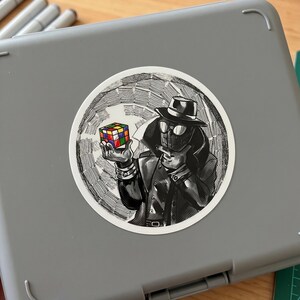 May include: A black and white sticker featuring a stylized illustration of a figure in a trench coat and fedora holding a Rubik's Cube. The cube has colorful squares. The sticker is round and set against a gray background.
