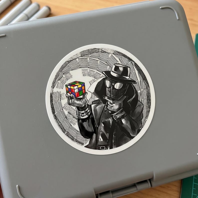 May include: A black and white sticker featuring a stylized illustration of a figure in a trench coat and fedora holding a Rubik's Cube. The cube has colorful squares. The sticker is round and set against a gray background.