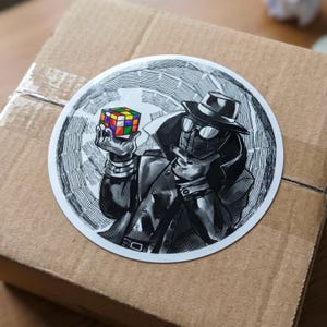 May include: A round sticker featuring a black and white illustration of a character in a fedora and trench coat, with a spiderweb design. The character is holding a colorful Rubik's Cube. The sticker is on a brown cardboard box.