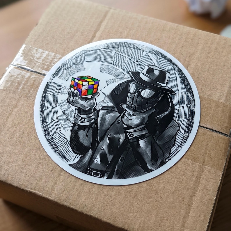May include: A round sticker featuring a black and white illustration of a character in a fedora and trench coat, with a spiderweb design. The character is holding a colorful Rubik's Cube. The sticker is on a brown cardboard box.