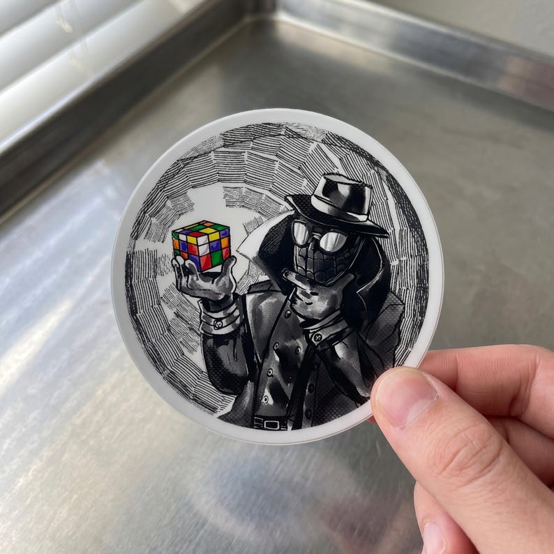 May include: A round sticker featuring a black and white illustration of a character in a fedora and trench coat, holding a colorful Rubik's Cube. The background has a radial pattern.