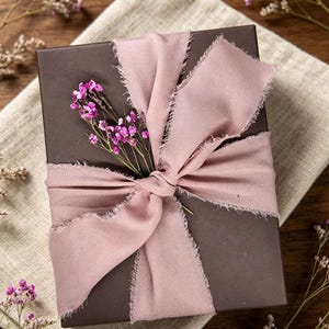 Può includere: Una scatola regalo rettangolare marrone scuro avvolta da un ampio nastro in tessuto rosa polvere con bordi sfrangiati. Un piccolo rametto di fiori viola essiccati è inserito sotto il nodo del nastro, al centro della parte superiore.