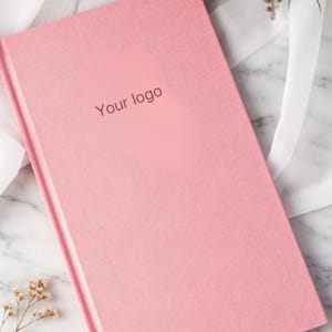 May include: A rectangular pink hardcover book with a textured finish. The front cover features the text Your logo printed in a dark pink font. The book is positioned at an angle, showcasing its solid color and clean, simple design aesthetic.