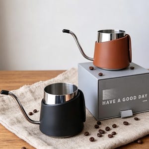 Puede incluir: Dos teteras de café de acero inoxidable con boquillas largas y finas, con fundas protectoras en negro y marrón. Hay una caja gris con el texto HAVE A GOOD DAY, junto con granos de café esparcidos sobre una superficie de tela texturizada.