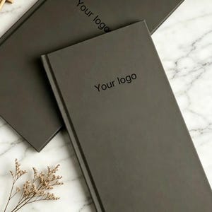 May include: Two rectangular dark gray notebooks with a matte finish, each featuring the text Your logo printed in a simple black sans-serif font on the front cover. The notebooks are positioned at an angle, showcasing their clean, solid-colored design.