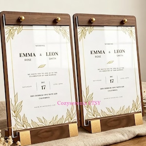 May include: Two vertical wedding invitation signs held in dark wood frames with gold metal accents. The white cards feature gold leaf illustrations, black text reading Emma & Leon Rose Smith, and event details for Saturday, December 17th.