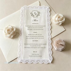 May include: A rectangular off-white fabric menu card with a delicate white lace border, featuring the text Menu, Antipasto, Primo, Secondo, and Dolce, accompanied by two cream-colored fabric roses and a gold ring in a velvet ring box.