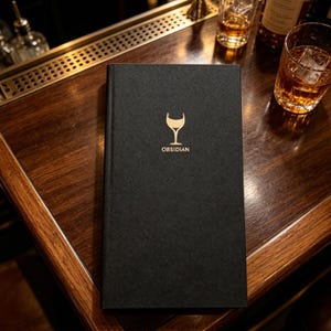 May include: A rectangular black menu cover with a textured surface. Centered on the front is a gold-coloured graphic of a wine glass above the word OBSIDIAN printed in gold capital letters. The cover has a clean, vertical spine on the left side.