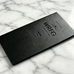 以下が含まれることがあります： 質感のある表面を持つ長方形の黒いメニューカバー。表面には「WRFG」、「TRATTORIA & PIZZERIA」、「SINCE 2005」、「MENU 2022」というエンボス加工の文字が施されています。デザインはカバーの中央に配置され、すっきりとしたプロフェッショナルな外観です。