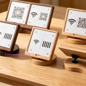 Puede incluir: Una colección de soportes de madera para letreros con inserciones de acrílico blanco que muestran símbolos Wi-Fi, códigos QR e iconos de líneas verticales impresos en negro. Los letreros están montados sobre bases circulares de madera.