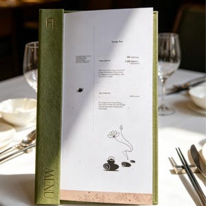 Puede incluir: Una carpeta de menú vertical con una cubierta de color verde oliva texturizado que presenta la palabra MENU y las letras FL en relieve. La página interior blanca muestra el texto Green Tea, precios y un dibujo lineal negro de una tetera y loto.