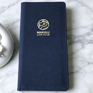 May include: A rectangular navy blue wallet with a smooth, matte finish and stitched edges. Centred on the front is a gold-coloured circular logo featuring a lion profile and a small crown, with the text MAKULU LION CLUB printed below in gold.