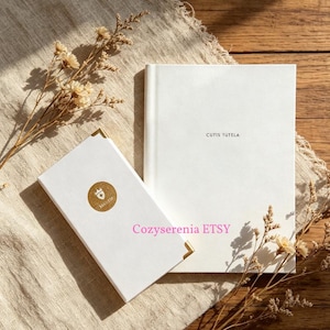 May include: A white rectangular box with gold corner accents and a circular gold sticker featuring a crown logo, alongside a white hardcover book with the text CUTIS TUTELA printed in black on the front cover, accompanied by dried flowers.