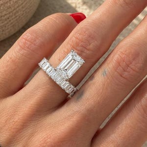 May include: A silver-toned ring featuring a large emerald-cut center stone set vertically, accompanied by a band adorned with a row of smaller, rectangular-cut stones that extend horizontally across the finger, creating an asymmetrical design.