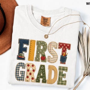 May include: A white t-shirt featuring the text FIRST GRADE in large, multi-patterned, fabric-style letters. The letters include designs like plaid, polka dots, notebook paper, and stars, with small graphics of books, an apple, and pencils.