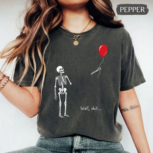 Peut inclure: Un t-shirt gris anthracite orné d'un motif de squelette blanc, d'un ballon rouge tenu par une main squelettique et du texte "Well, shit..." imprimé en blanc. Le t-shirt présente un col rond et des manches courtes, avec une coupe décontractée.