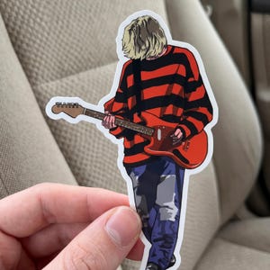 May include: A die-cut sticker featuring a stylized illustration of a person with blonde hair wearing a red and black horizontal striped long-sleeve shirt and blue jeans, playing a solid-body electric guitar with a red finish and a brown neck.
