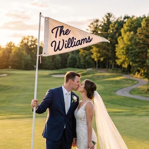 May include: A couple kissing on a golf course, with a flag that says "The Williams". The bride wears a white lace wedding dress and veil, holding a bouquet. The groom wears a navy suit. The scene is bathed in warm sunlight.
