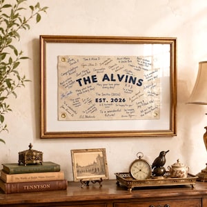 以下が含まれることがあります： THE ALVINS EST. 2026という濃い青色の文字が中央にあり、その周囲を黒いインクの手書きメッセージや署名が囲んでいる、額装された長方形の布製バナー。バナーの四隅には金色の金属製ハトメが付いています。