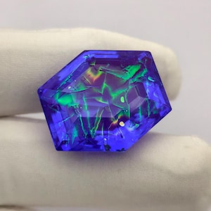 May include: A hexagonal gemstone with a deep blue colour and internal fractures. The gem displays flashes of green, yellow, and purple. It is held against a white background.