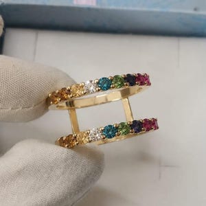 May include: A gold double-band ring featuring two parallel rows of small, round, multi-colored gemstones. The stones are arranged in a gradient pattern including yellow, white, blue, green, purple, and pink hues set into the gold metal bands.