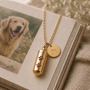 May include: A gold-toned metal pendant necklace featuring a cylindrical urn charm with two cut-out paw prints and a circular tag engraved with the name Max and the date August 7, 2026, suspended from a delicate matching gold-toned chain link necklace.