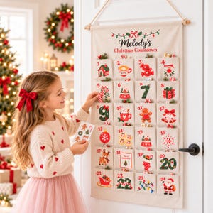 May include: A beige fabric wall-hanging advent calendar with the text Melody's Christmas Countdown. It features twenty-four numbered pockets, each decorated with colourful holiday illustrations including a snowman, gingerbread man, and Santa Claus.