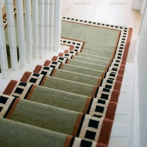 Puede incluir: Una alfombra para escaleras con un centro verde salvia, un borde decorativo en color naranja terracota y un patrón geométrico repetitivo de cuadrados negros y blancos, instalada sobre escalones de madera blanca con barandilla.