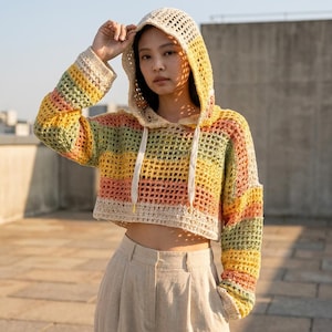 May include: A cropped, long-sleeve hooded sweater featuring a loose, open-knit crochet pattern with horizontal stripes in shades of yellow, orange, green, and cream. The garment includes a hood with two white drawstring cords hanging down the front.