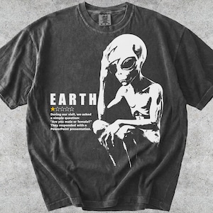Może przedstawiać: Ciemnoszary t-shirt z krótkim rękawem z białą grafiką przedstawiającą kosmitę trzymającego rękę na głowie. Tekst głosi: EARTH, jedna żółta gwiazdka oraz: During our visit, we asked a simple question: Are you male or female? They responded with a PowerPoint presentation.
