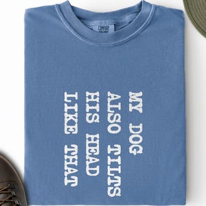 May include: A blue short-sleeved t-shirt featuring the white text "MY DOG ALSO TILTS HIS HEAD LIKE THAT" printed vertically. The shirt has a crew neckline and a Comfort Colors brand label visible on the inside of the collar area.