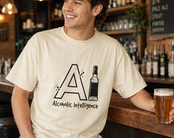 May include: A cream-colored short-sleeve t-shirt featuring a graphic of the letter A with pixelated accents and a bottle illustration, accompanied by the text Alcoholic Intelligence printed in a black sans-serif font on the front chest area.