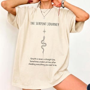 Puede incluir: Una camiseta beige de manga corta con un gráfico negro de una serpiente y una estrella. El texto dice THE SERPENT JOURNEY y Growth is never a straight line. Sometimes wisdom arrives after shedding everything you used to be.