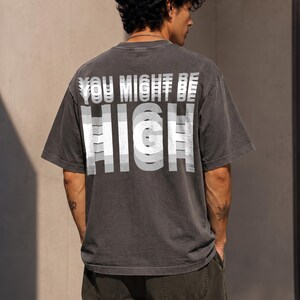May include: A charcoal gray short-sleeve t-shirt featuring a white graphic print on the back. The text reads YOU MIGHT BE HIGH in a layered, motion-blur style font. The shirt has a crew neckline and a relaxed fit, constructed from cotton fabric.