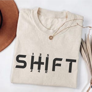 Könnte beinhalten: Ein cremefarbenes Kurzarm-T-Shirt mit dem Wort SHIFT in schwarzen Großbuchstaben, wobei der Buchstabe H durch ein manuelles Schaltgetriebemuster mit 1, 3, 5, 2, 4 und R ersetzt ist. Das T-Shirt hat einen Rundhalsausschnitt.