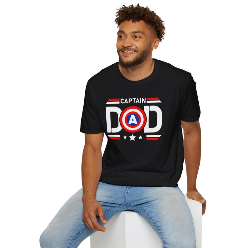 May include: Black t-shirt with the text "CAPTAIN DAD" in white, red, and blue. The design includes a shield with the letter "A" inside a red circle, and three white stars below. The shirt is a crew neck.