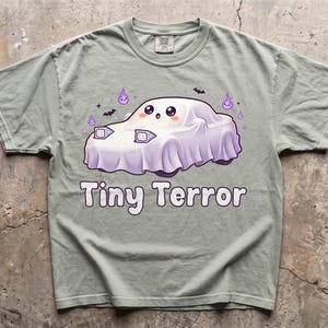 May include: A sage green t-shirt featuring a graphic of a car covered by a white ghost sheet with pop-up headlights, purple flames, and bats. The text Tiny Terror is printed in white below the car illustration. The shirt has a crew neckline.