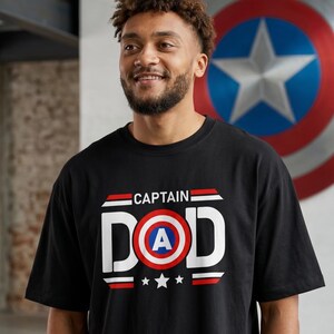 May include: Black t-shirt with the text "CAPTAIN DAD" in white, red, and blue, featuring a shield graphic with a central "A" and three stars. The shirt is worn by a person with curly hair and a beard.