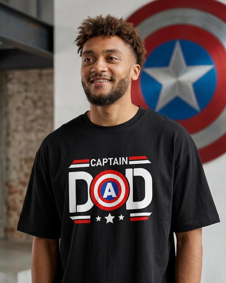 May include: Black t-shirt with the text "CAPTAIN DAD" in white, red, and blue, featuring a shield graphic with a central "A" and three stars. The shirt is worn by a person with curly hair and a beard.