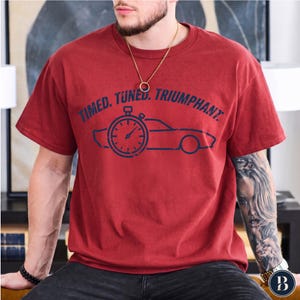 Pode incluir: Uma t-shirt vermelha de manga curta com uma impressão gráfica azul-escura de um cronómetro e a silhueta de um carro. O texto "TIMED. TUNED. TRIUMPHANT." está impresso em azul-escuro acima do gráfico. A t-shirt tem um decote redondo.