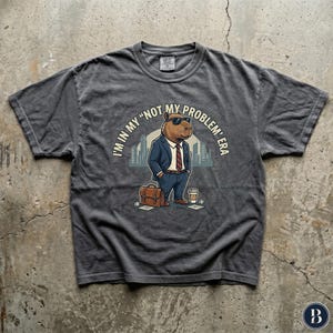 May include: A charcoal gray short-sleeve t-shirt featuring a graphic of a capybara wearing a suit, tie, and sunglasses. The capybara holds a briefcase and stands near a coffee cup. The text reads, I'M IN MY "NOT MY PROBLEM" ERA.