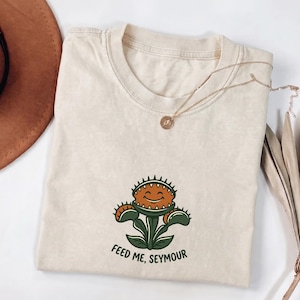 May include: A beige crew neck t-shirt featuring a graphic of a smiling Venus flytrap plant with green leaves and a brown mouth. The text "FEED ME, SEYMOUR" is printed in black below the plant graphic. A gold-toned necklace rests on the neckline.