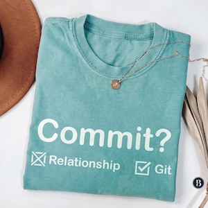 Può includere: Una t-shirt girocollo color verde acqua con il testo bianco "Commit?" sopra due caselle di controllo. La casella a sinistra ha una X accanto alla parola "Relationship" e quella a destra un segno di spunta accanto alla parola "Git". Una collana d'oro.