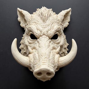 Puede incluir: Una máscara de pared de color crema que representa la cabeza de un jabalí. La escultura presenta una textura de pelaje detallada, orejas puntiagudas, cuencas oculares profundas y dos colmillos curvos prominentes a ambos lados del hocico.